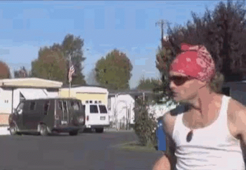 A Man Wearing A Red Bandana And Sunglasses Is Walking Down A Street In A Trailer Park . GIF