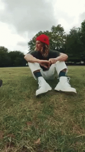 A Man Wearing A Red Beanie And White Boots Is Sitting On The Grass . GIF
