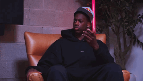 A Man Wearing A Red Bull Hat Sits In A Chair Looking At His Phone GIF