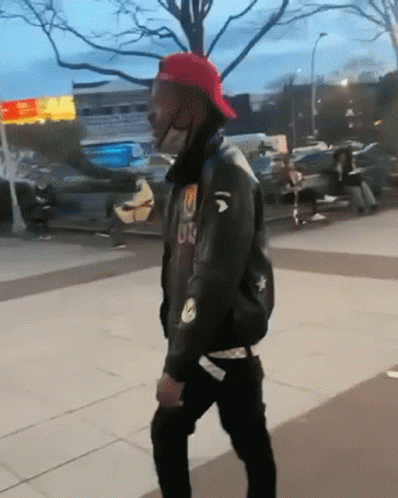 A Man Wearing A Red Hat And A Black Jacket With The Letter U On It Is Walking Down A Sidewalk GIF