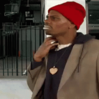 A Man Wearing A Red Hat And A Heart Necklace Is Standing In Front Of A Fence . GIF
