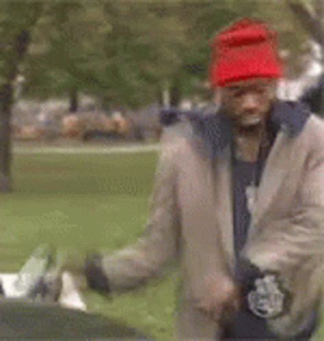A Man Wearing A Red Hat And A Tan Jacket Is Standing In A Park . GIF
