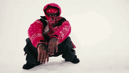a man wearing a red jacket and a bandana on his face is squatting down in the snow .