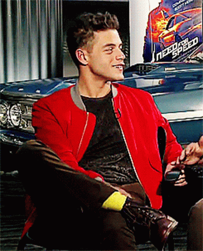 A Man Wearing A Red Jacket Sits In Front Of A Need For Speed Poster GIF