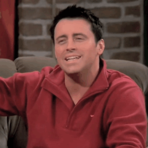 A Man Wearing A Red Nike Sweater Smiles While Sitting On A Couch GIF