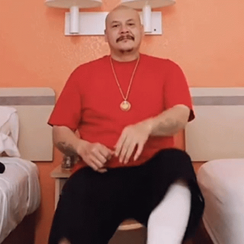 A Man Wearing A Red Shirt And A Gold Necklace Is Sitting On A Chair GIF