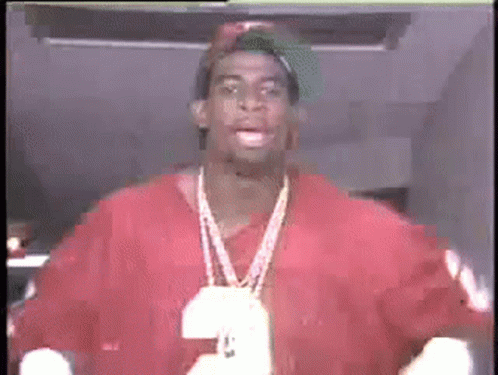 A Man Wearing A Red Shirt And A Necklace With A Pendant That Has The Letter L On It GIF