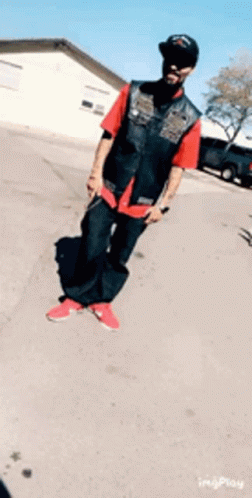 A Man Wearing A Red Shirt And Black Vest Is Standing On A Street GIF
