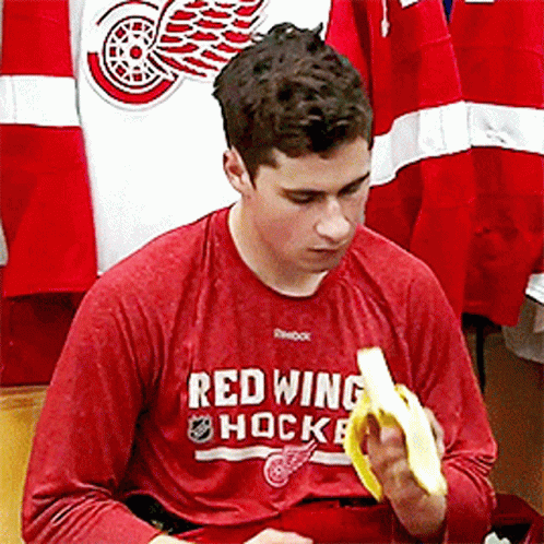 A Man Wearing A Red Shirt That Says Red Wing Hockey Is Eating A Banana GIF