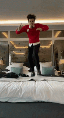 A Man Wearing A Red Sweater Is Jumping On A Bed GIF