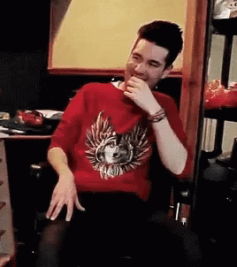 A Man Wearing A Red Sweater With A Wolf On It Is Sitting In A Chair And Laughing . GIF