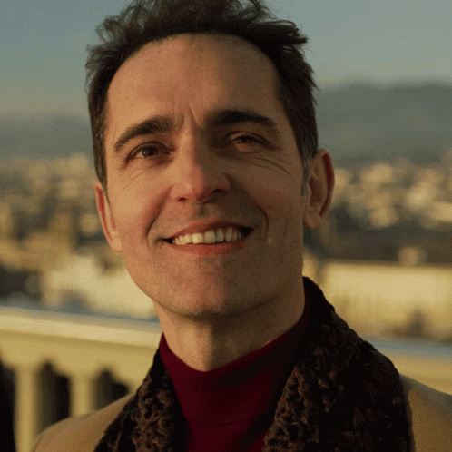 A Man Wearing A Red Turtleneck And A Brown Scarf Smiles For The Camera GIF