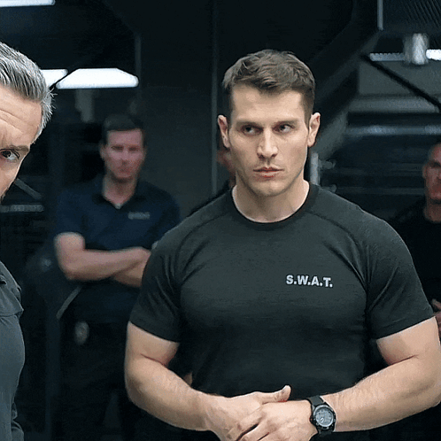 A Man Wearing A S.w.a.t. T-Shirt Stands In Front Of Another Man GIF