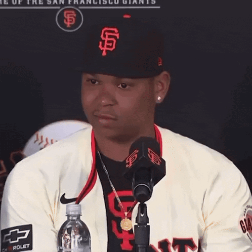 A Man Wearing A San Francisco Giants Hat Stands In Front Of A Microphone Holding A Bottle Of Water GIF