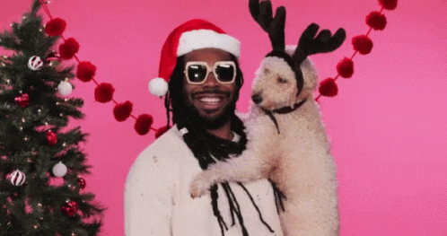 A Man Wearing A Santa Hat And Sunglasses Holds A Dog With Reindeer Antlers GIF