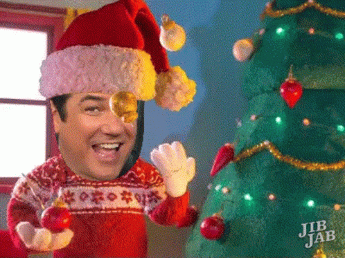 A Man Wearing A Santa Hat And Sweater Is Holding A Christmas Ball In Front Of A Christmas Tree GIF