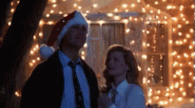 A Man Wearing A Santa Hat Stands Next To A Woman In Front Of Christmas Lights GIF