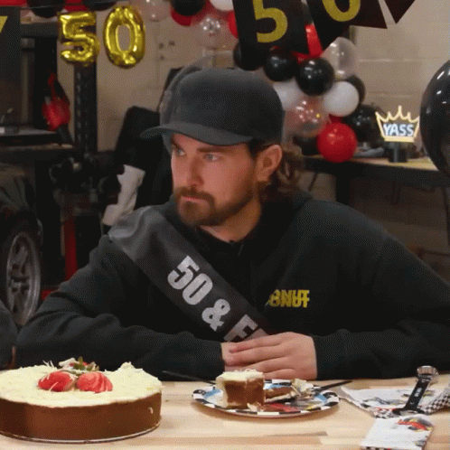 A Man Wearing A Sash That Says 50 & F Sits At A Table With A Cake GIF