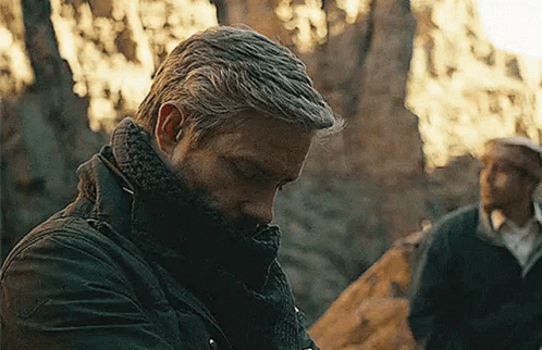 A Man Wearing A Scarf Around His Neck Is Standing In The Woods Looking Down . GIF