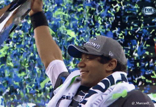 A Man Wearing A Seahawks Hat Is Holding Up A Trophy GIF