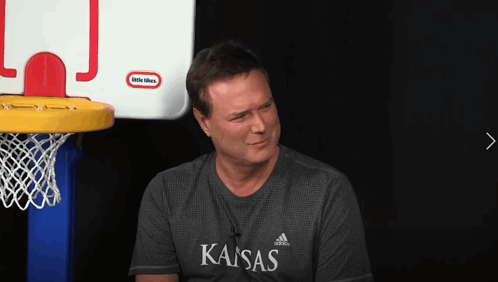 A Man Wearing A Shirt That Says Kansas Sits In Front Of A Little Tikes Basketball Hoop GIF