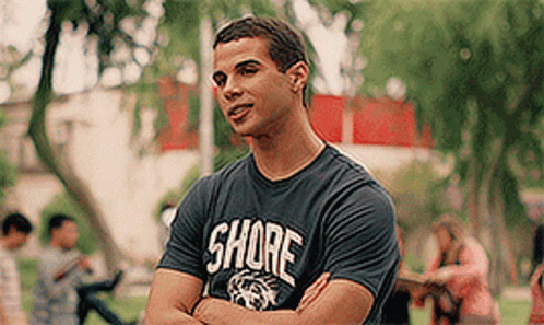 A Man Wearing A Shore T-Shirt Is Standing In A Park With His Arms Crossed GIF