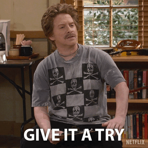 A Man Wearing A Skull And Crossbones Shirt Is Sitting In Front Of A Bookshelf And Says Give It A Try GIF