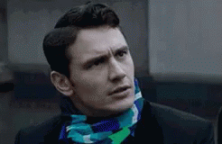A Man Wearing A Suit And A Scarf Around His Neck Is Looking At The Camera . GIF