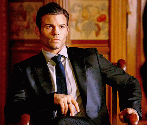 A Man Wearing A Suit And Tie Is Sitting In A Chair . GIF