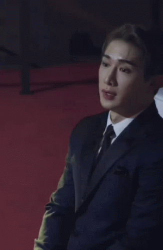 A Man Wearing A Suit And Tie Is Sitting On A Red Carpet . GIF