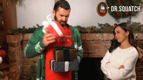 A Man Wearing A Sweater That Says Christmas Y'all Is Holding A Stack Of Gifts GIF