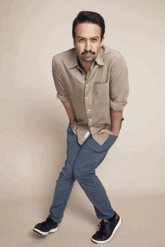 A Man Wearing A Tan Shirt And Blue Pants Is Standing With His Hands In His Pockets GIF