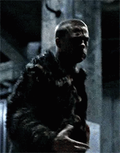 A Man Wearing A Tank Top And A Leather Jacket Is Sitting Down With His Arms Outstretched In A Dark Room . GIF