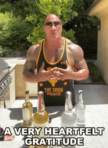 A Man Wearing A Tank Top That Says The Iron Paradise Stands In Front Of Bottles Of Alcohol GIF