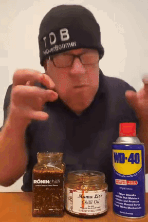 A Man Wearing A Tdb Hat Holds A Can Of Wd-40 And A Jar Of Mama Liz 'S Chilli Oil GIF