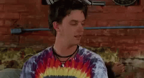 A Man Wearing A Tie Dye Shirt And A Choker Is Sitting On A Couch In Front Of A Brick Wall . GIF