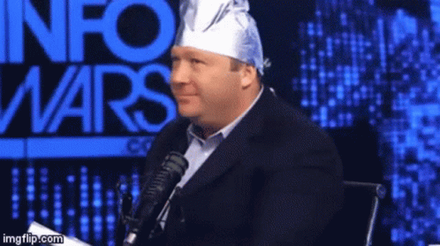 A Man Wearing A Tin Foil Hat Is Sitting In Front Of A Microphone In Front Of An Info Wars Sign GIF