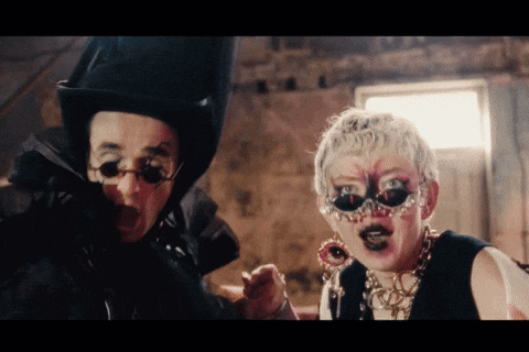 A Man Wearing A Top Hat And Glasses Stands Next To A Woman Wearing A Mask And Earrings GIF