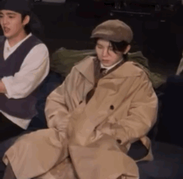 A Man Wearing A Trench Coat And Hat Is Sitting On A Chair . GIF