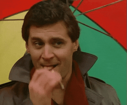A Man Wearing A Trench Coat And Scarf Is Biting A Cigarette While Holding A Colorful Umbrella GIF