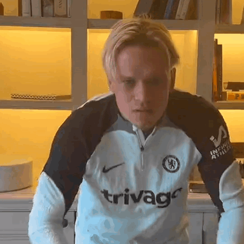 A Man Wearing A Trivago Jersey Stands In Front Of A Bookshelf GIF
