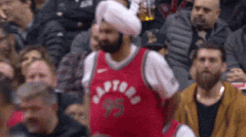 A Man Wearing A Turban And A Raptors Jersey Sits In The Crowd GIF
