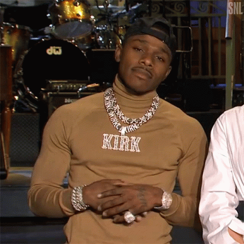 A Man Wearing A Turtleneck And A Necklace With The Name Kirk On It Is Standing In Front Of A Drum Set . GIF