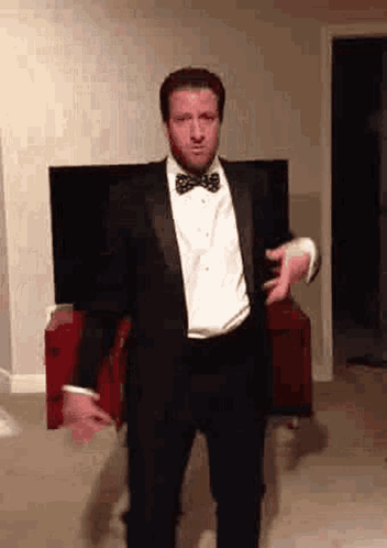 A Man Wearing A Tuxedo And Bow Tie Is Dancing In A Living Room . GIF