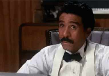 A Man Wearing A Tuxedo And Bow Tie Is Sitting At A Desk . GIF