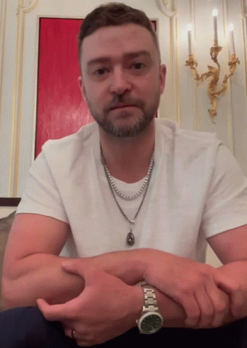 A Man Wearing A Watch And A Necklace Is Sitting With His Hands Folded GIF