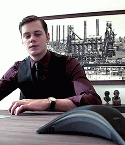 A Man Wearing A Watch Sits At A Desk In Front Of A Picture GIF
