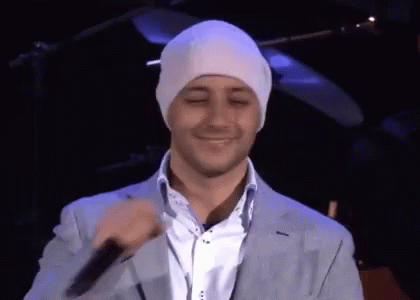 A Man Wearing A White Beanie And A Suit Is Smiling And Holding A Microphone GIF