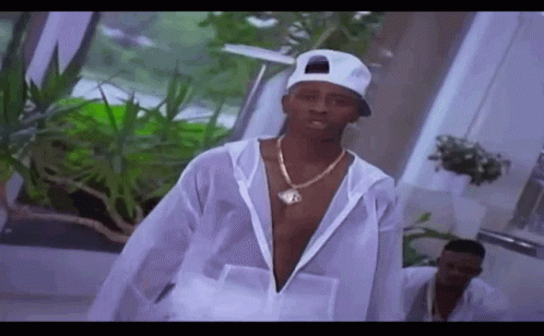 A Man Wearing A White Hat And A Necklace Is Standing In A Room With Plants . GIF