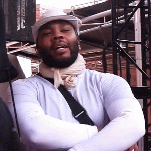 A Man Wearing A White Hat And A Scarf Around His Neck Is Standing With His Arms Crossed GIF